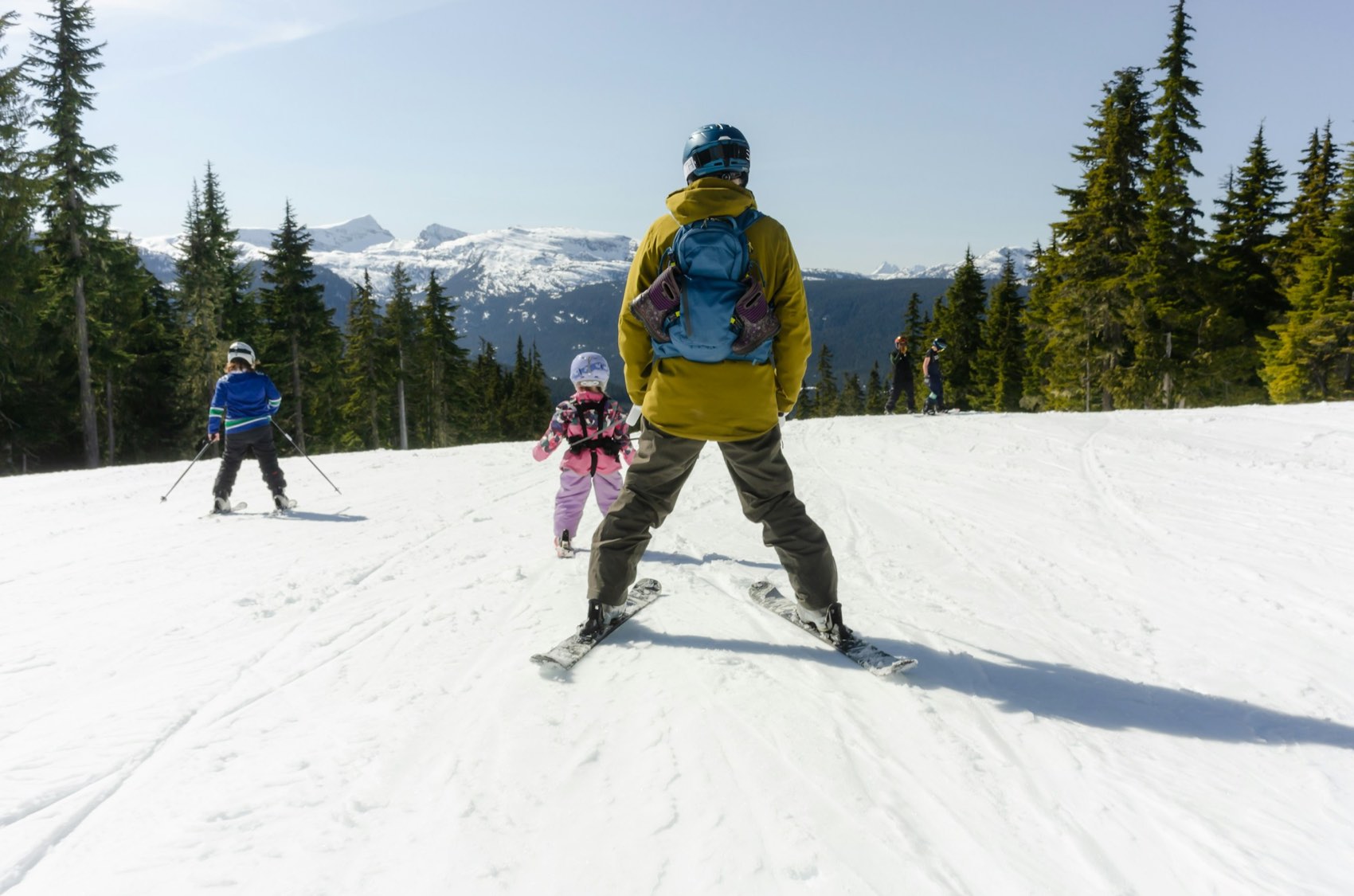 An adult and two children on skis at the top of a groomed run, glaciated coastal peaks behind them