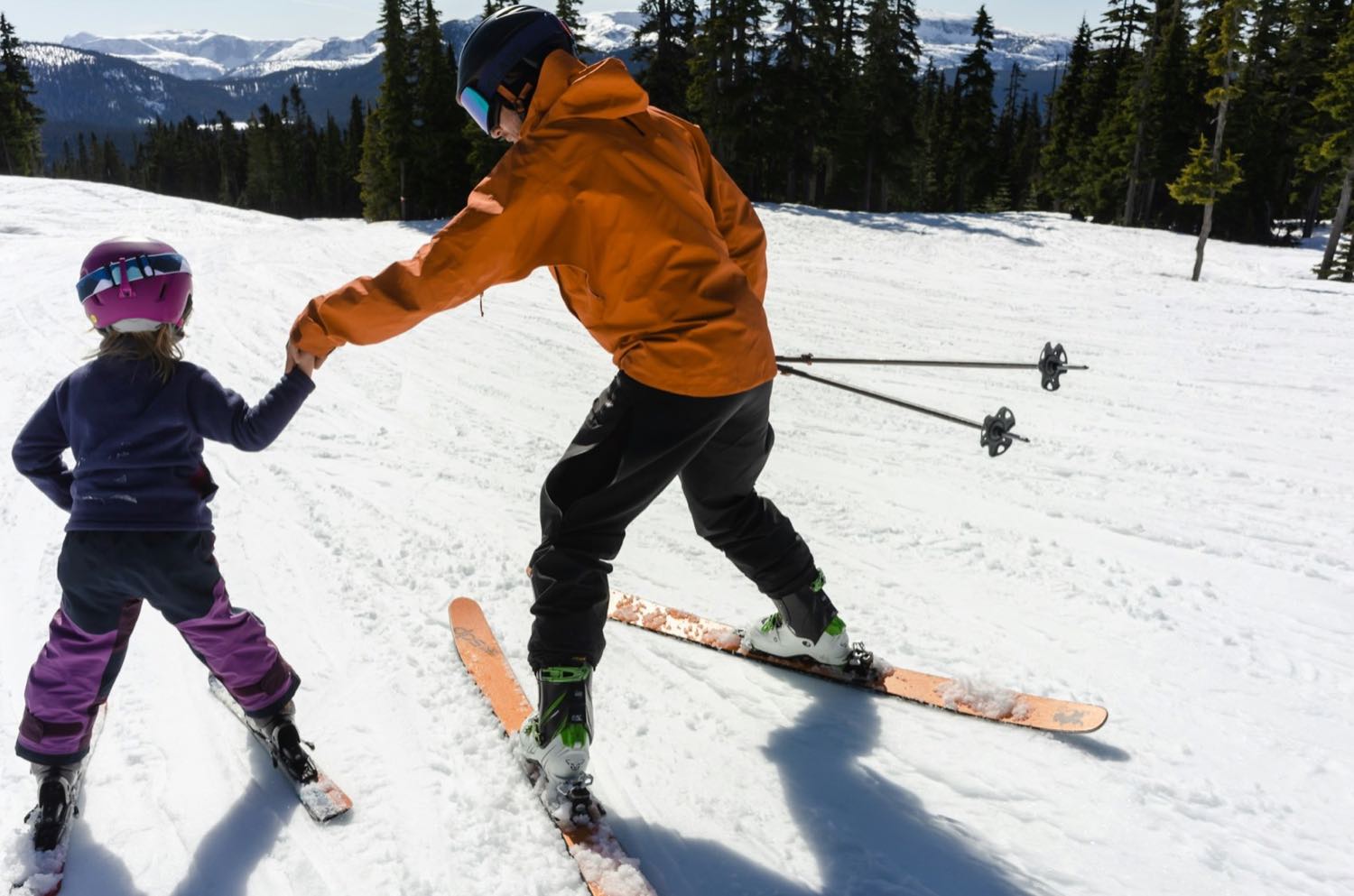 An adult in an orange jacket holding a young skier's hand on a groomed run, snowy coastal peaks behind