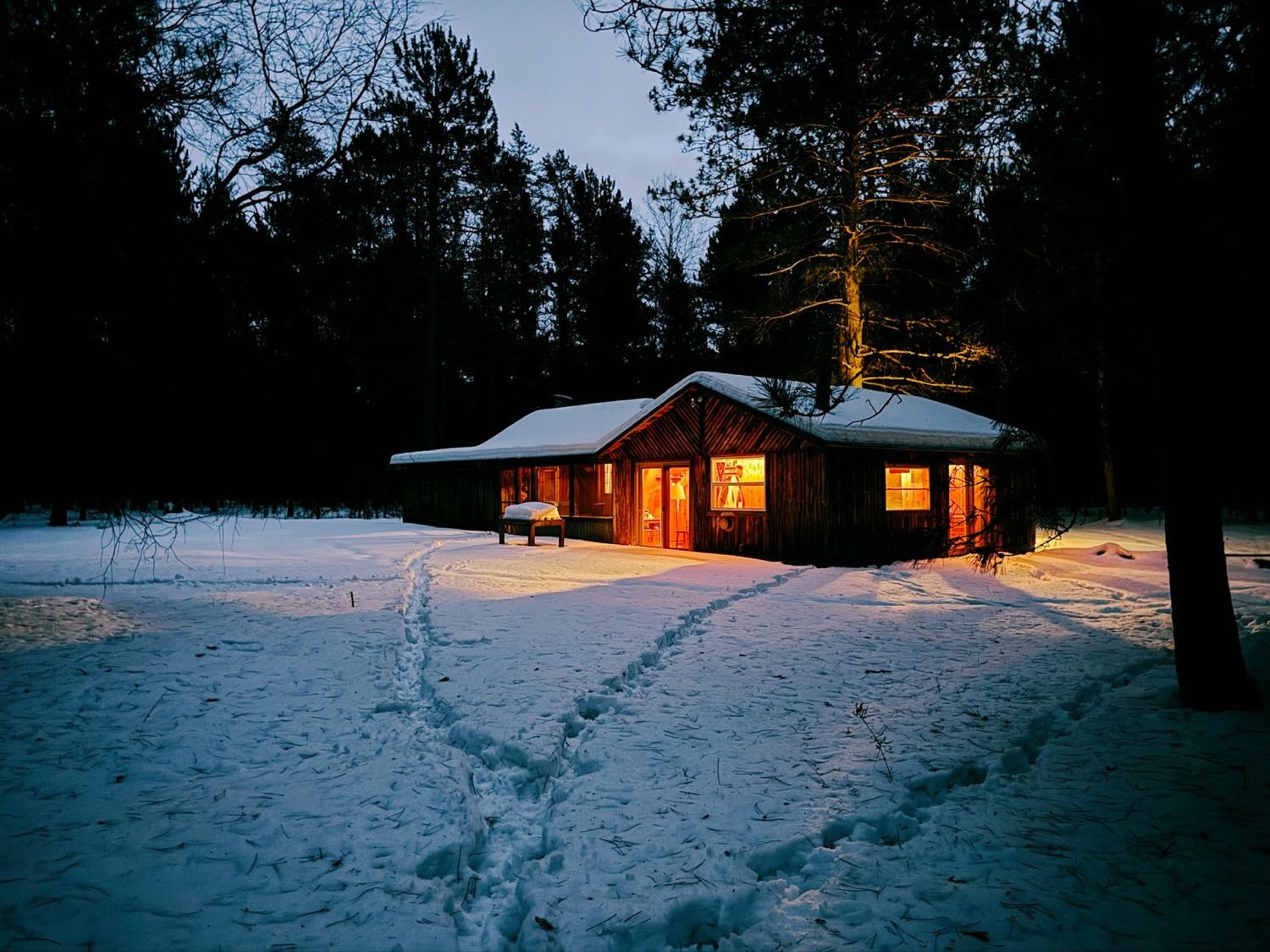 A low timber building with warmly lit windows sitting in deep snow among tall trees at dusk