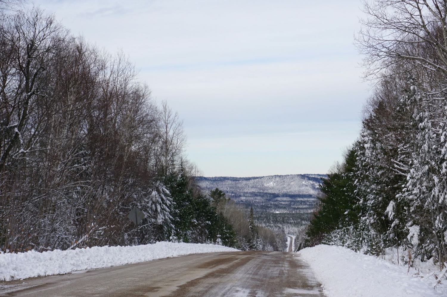 A snow-banked two-lane road descending through winter forest towards distant forested hills