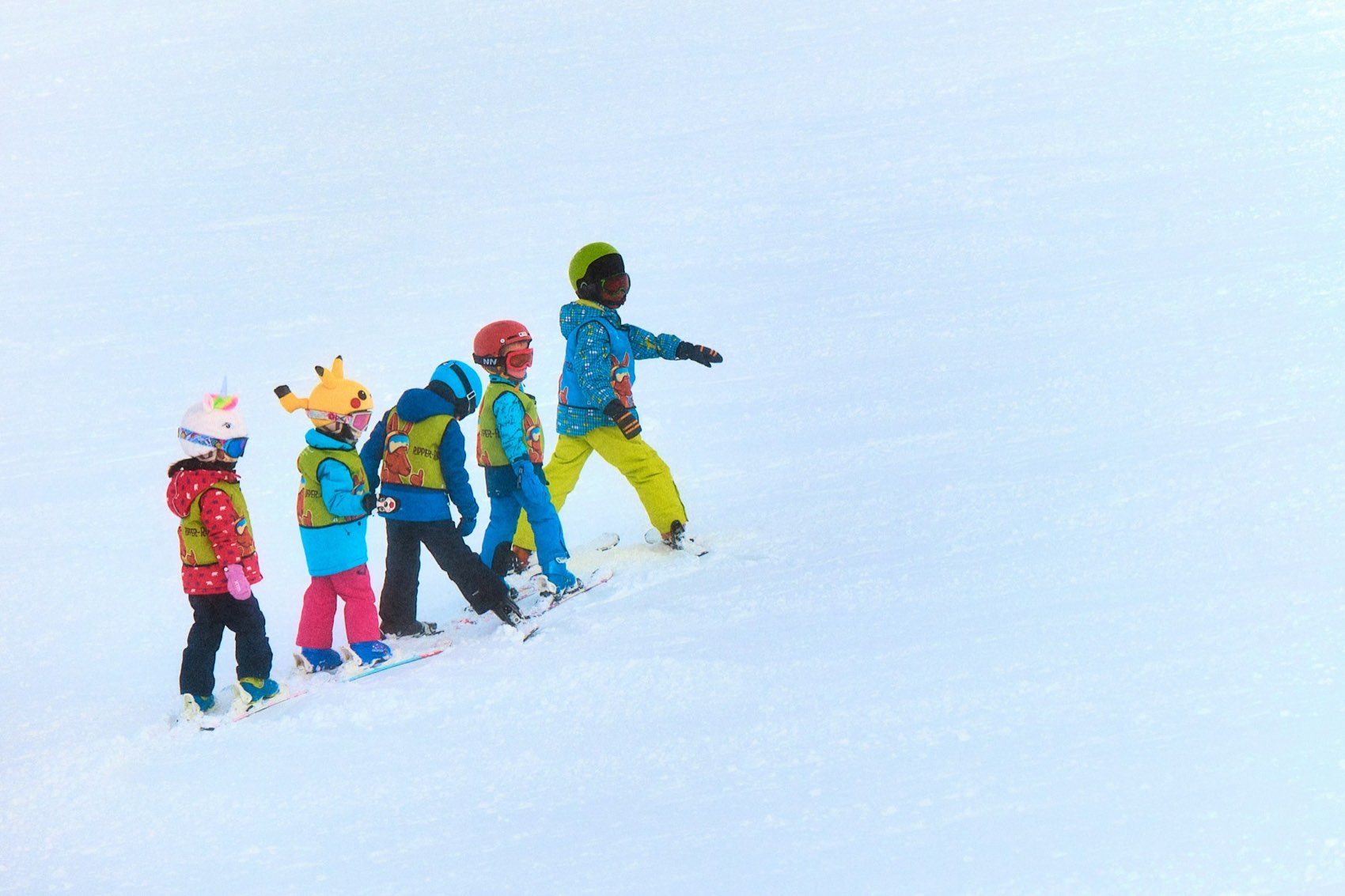 Five small children in bright helmets and ski-school bibs walking in a line across the snow